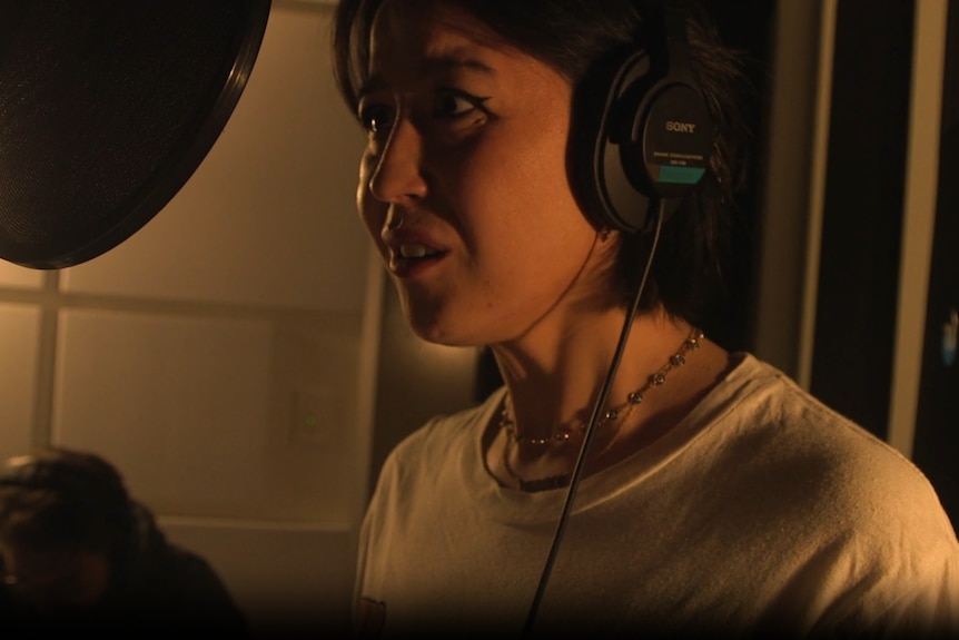 Nina Oyama with headphones on speaking into a mic in a darkened room