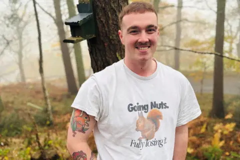 Joel Sangster is wearing a 'going nuts' T-shirt which features a squirrel, smiling at the camera, while standing in woods, and he also has a squirrel tattoo on his arm.
