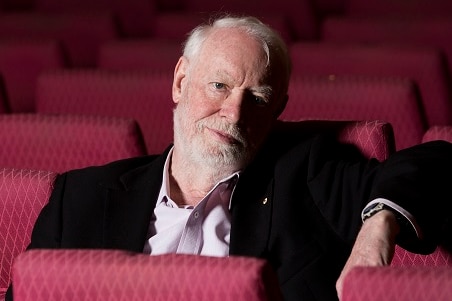 man with white hair and beard sitting alone in a theatre with red chairs