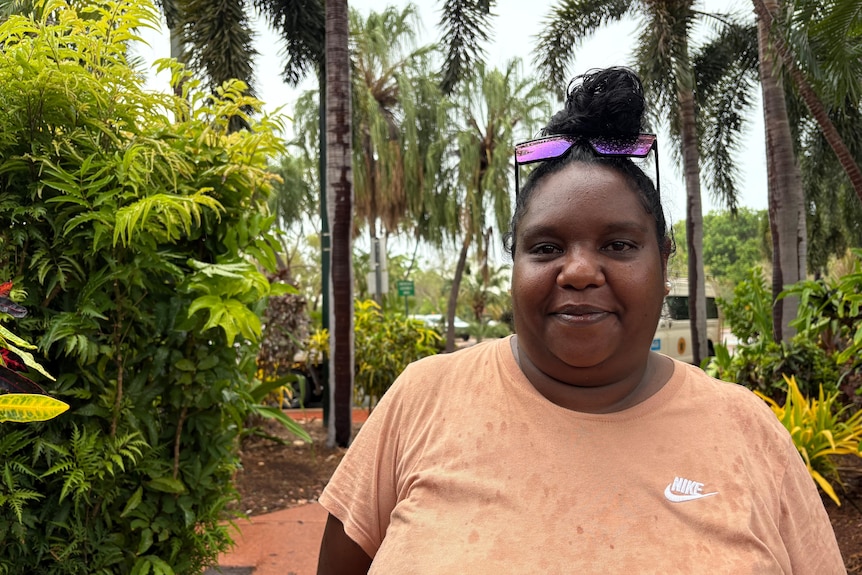 A woman wearing a peach coloured t shirt smiles at the camera with water droplets on her shirt and palm trees outside.