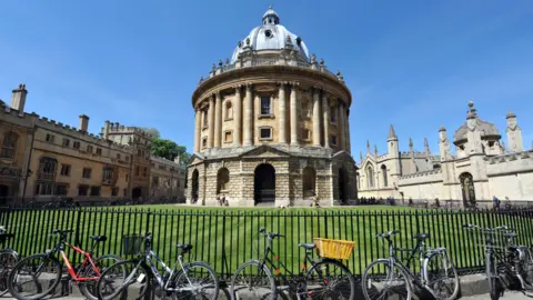 The Bodleian Library surrounded by a black metal fence and green lawn. Several bicycles are parked along the fence. Adjacent Gothic-style buildings with spires and arched windows are visible under a clear blue sky.