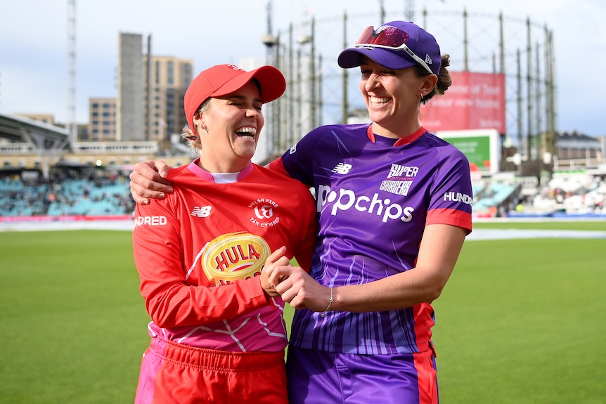 Alex Hartley and Kate Cross laugh with their arms around one another on a cricket field.