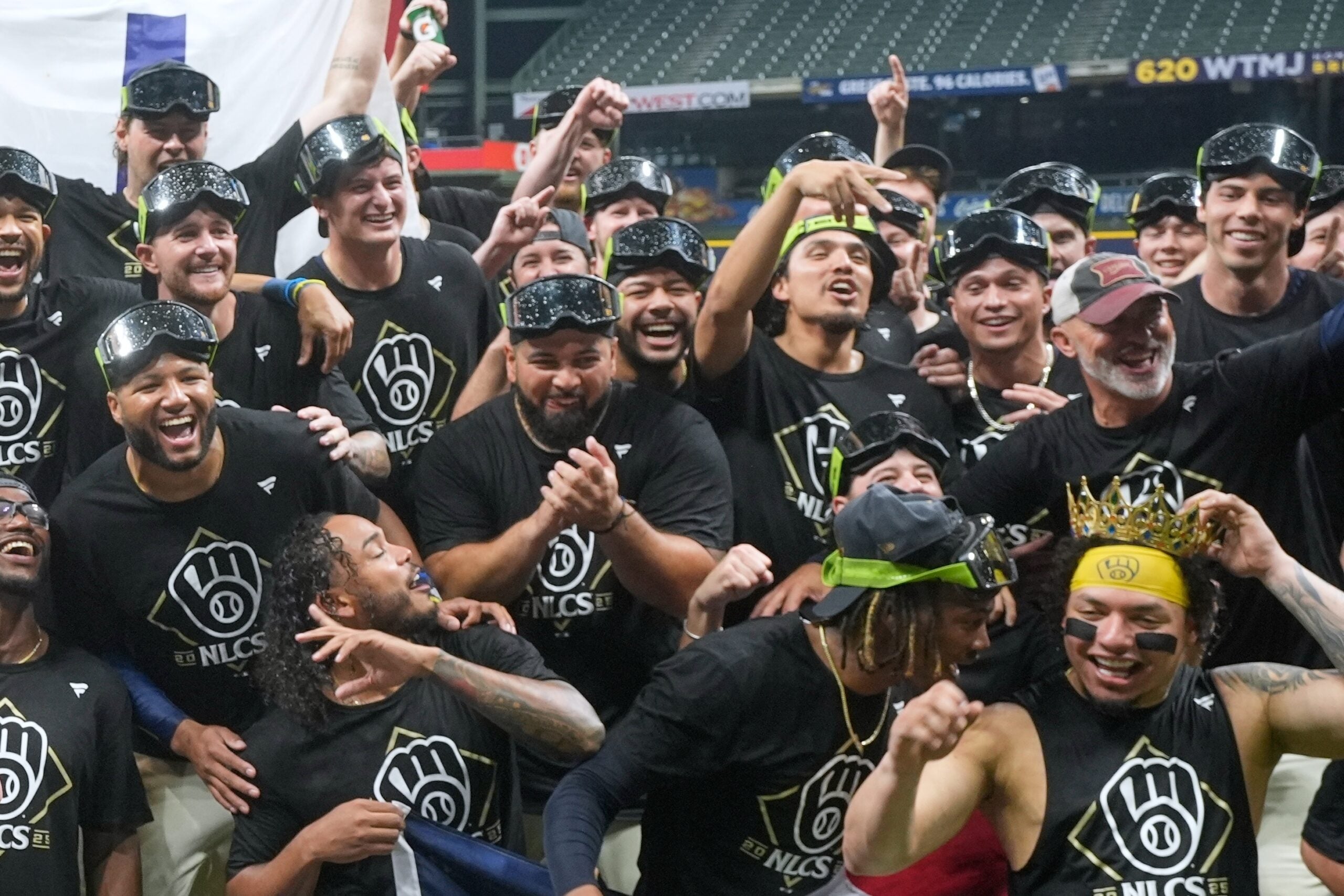 A group of baseball players and staff in matching shirts and goggles celebrate together on the field, smiling and posing for a team photo.