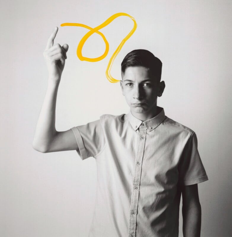 A young person in a buttoned shirt raises one hand, pointing upward, with a yellow squiggle drawn above their finger on a plain background.