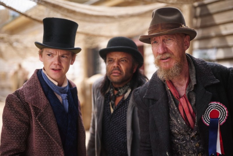 Three men dressed in period costumes, including top hats and vests, stand together in a western-themed setting, looking intently at something off-camera. Their expressions convey curiosity and concern, with a backdrop of rustic buildings and netting typical of a historical film scene.