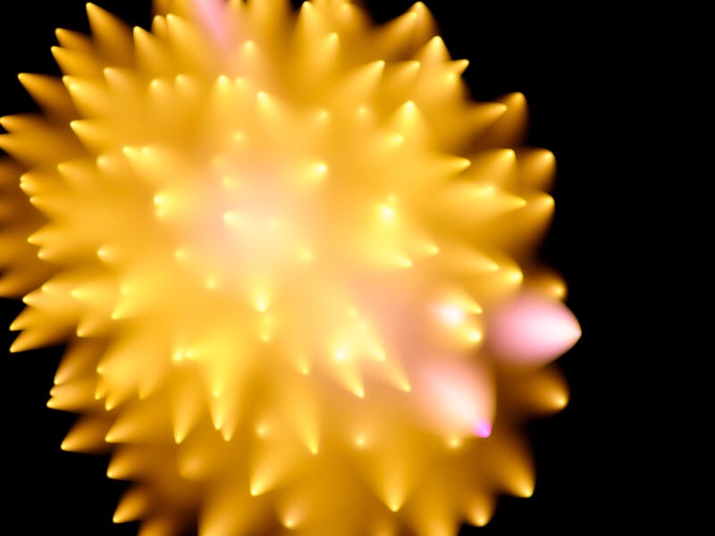 A bright, spiky, abstract yellow-orange shape with glowing tips set against a black background, giving a soft, radiant appearance with hints of pink on some of the spikes.