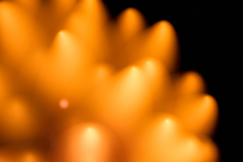 Abstract close-up image featuring blurred, glowing orange shapes with soft edges, creating a warm, radiant effect against a dark background. The shapes resemble petals or flame tips.