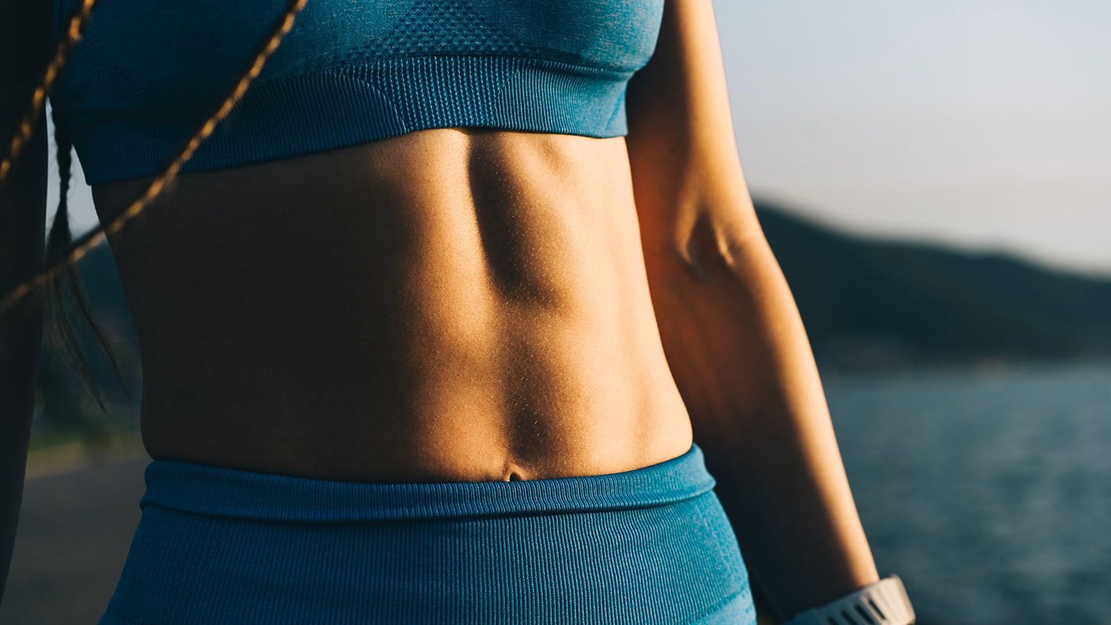 A close-up of a woman's athletic torso, showing her toned abs and a blue sports bra. Wearable health tracker on the hand. The golden hour sunlight highlights the muscles and fabric, creating a powerful image that represents fitness goals, body confidence, and strength.