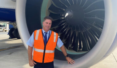 A man in an orange safety vest stands confidently beside a large aircraft jet engine on an airport tarmac, conveying a sense of professionalism. He uses V A's telehealth services.