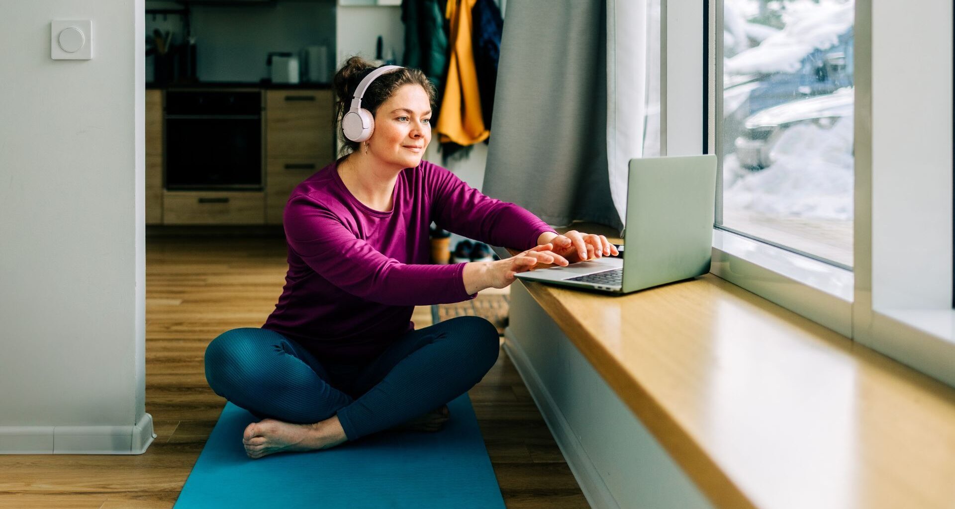 Woman setting up laptop to do low-impact cardio workout at home on yoga mat with snow outside window and wearing headphones