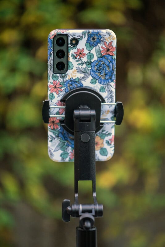 A smartphone with a colorful floral case is mounted on a black tripod, with a blurred background of green and yellow foliage.
