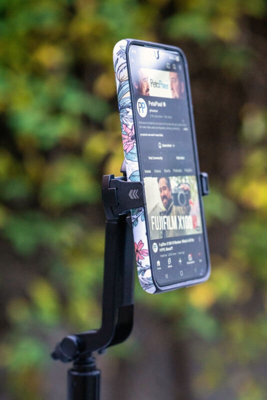 A smartphone with a floral-patterned case is mounted on a tripod, displaying a YouTube channel page. The background is blurred greenery.