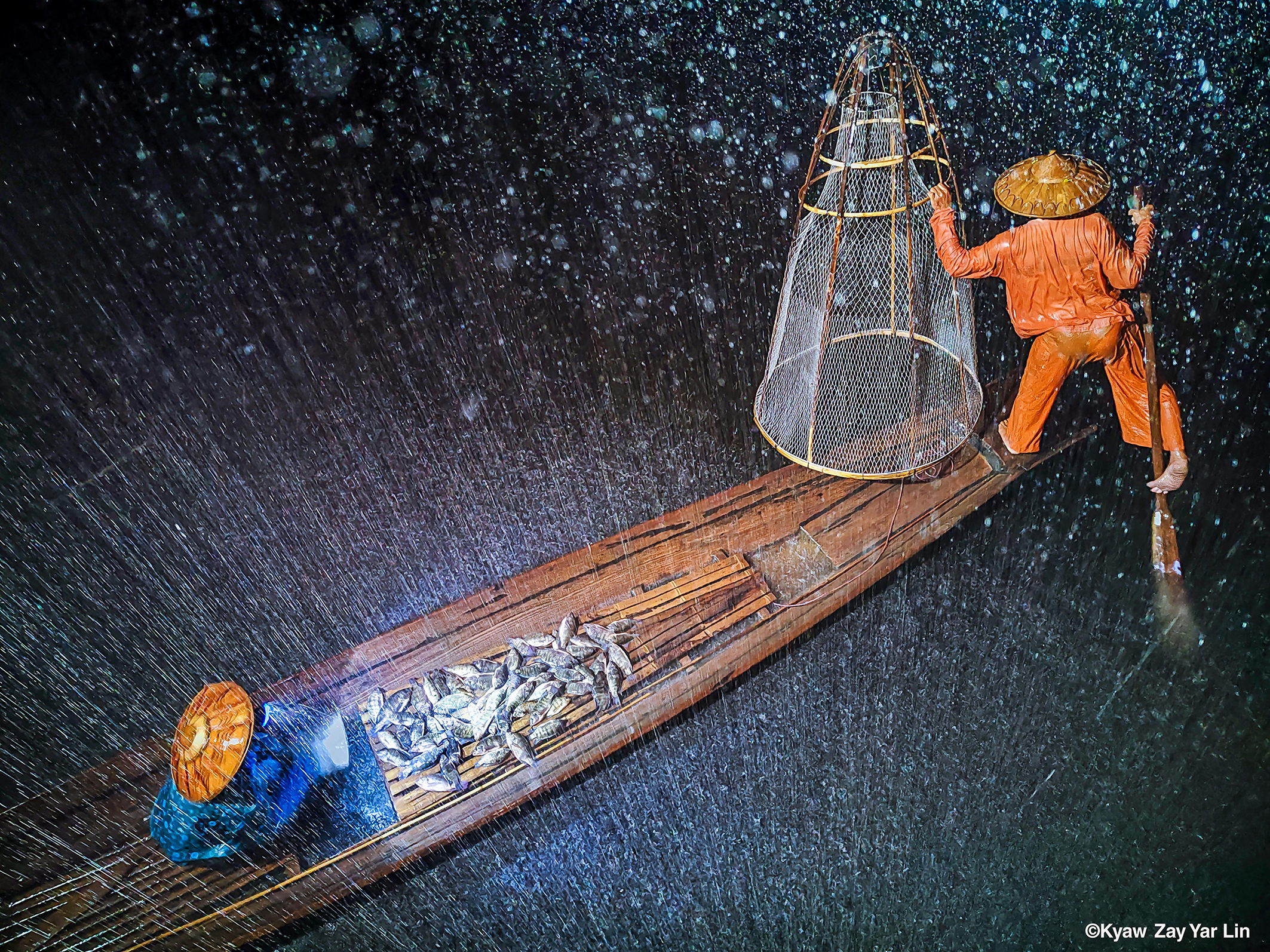 A fisherman in orange attire navigates a wooden boat, catching fish under heavy rain, with water splashing around him
