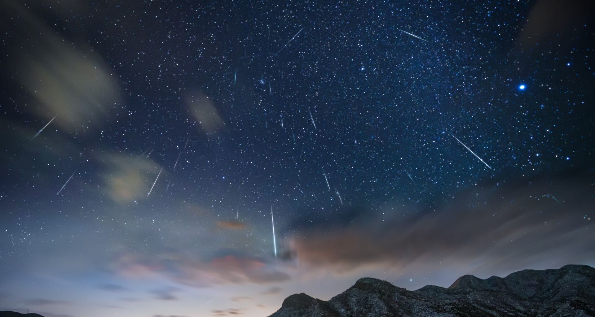 A series of white streaks of meteor showers are seen in a pale blue night sky over some mountain peaks at the bottom of the image