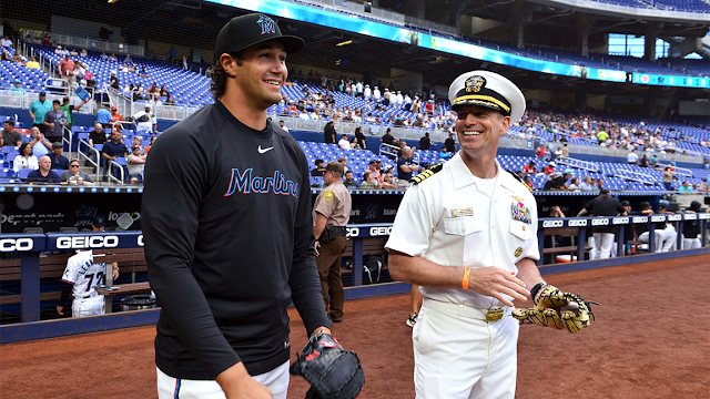 A smiling U.S. Navy officer in a white dress uniform stands on a professional baseball field holding a baseball glove, conversing with a Miami Marlins player dressed in a black warmup jersey and cap.