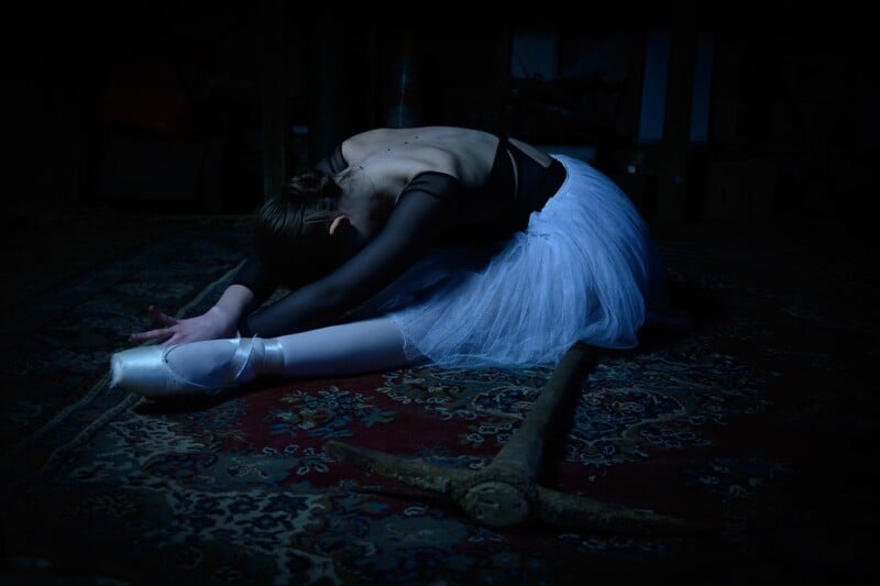 A ballerina in a white tutu and pointe shoes sits on a patterned rug, stretching forward with her arms extended, head down, and a pickaxe lying beside her in dim lighting.
