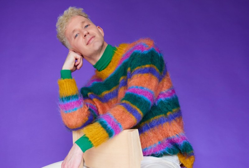 Young man with curly blonde hair wearing a colourful striped sweater, sitting on a wooden chair against a purple background, showcasing a relaxed and contemplative pose.