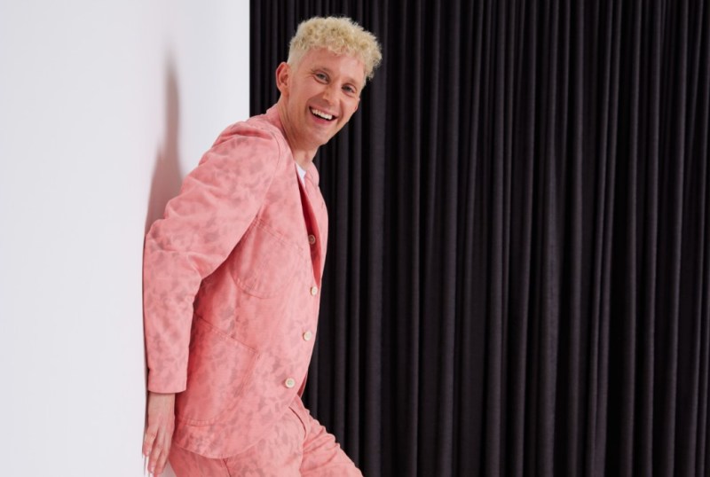 Smiling man in a pink suit posing playfully against a backdrop of black curtains, showcasing a modern fashion style.