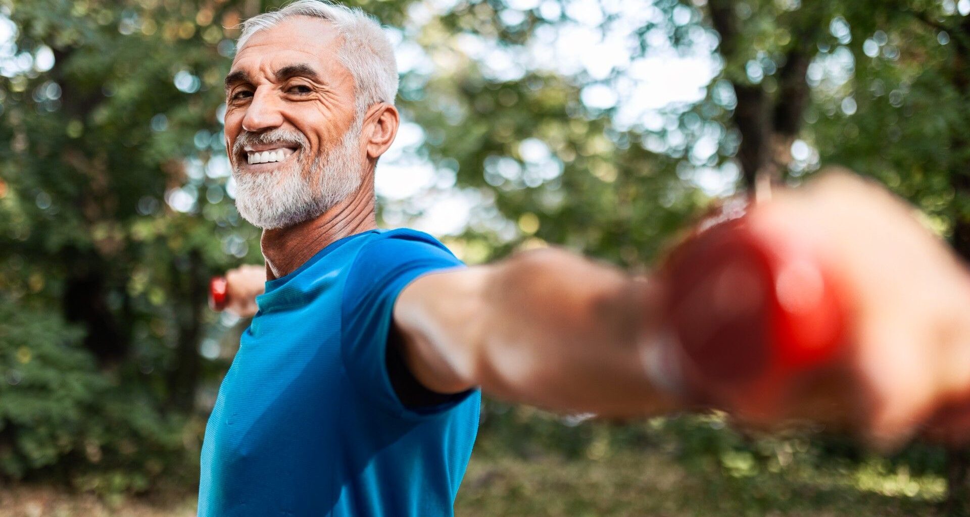 Older male in blue t-shirt holding dumbbells out at shoulder-height in a local park smiling