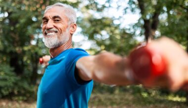 Older male in blue t-shirt holding dumbbells out at shoulder-height in a local park smiling