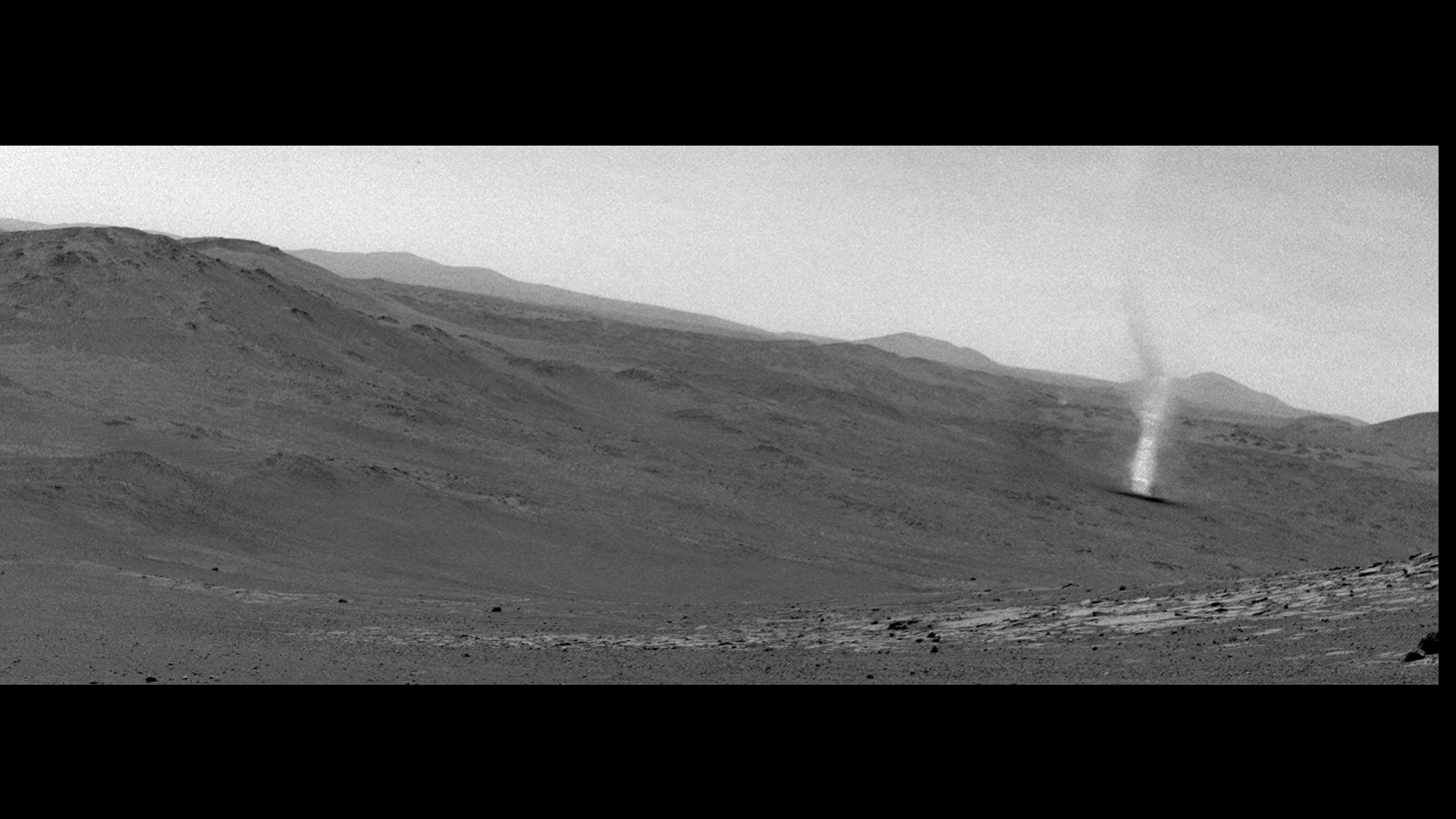 A grayscale still from a video showing a swirling cloud of dust along a dune on Mars