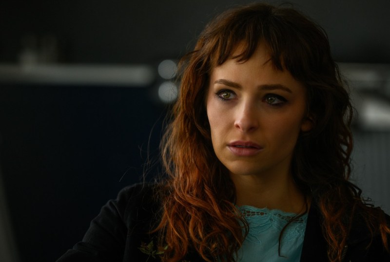 Close-up of a woman with wavy brown hair and green eyes, wearing a light blue top and a black jacket, displaying an expression of concern or contemplation. The background is softly blurred, focusing attention on her face. Ideal for articles on emotional expression in photography or character analysis in film.