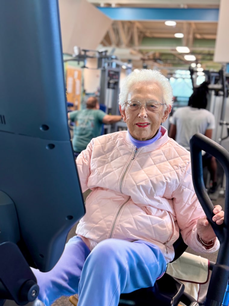 Ruth Lemay in a pink jacket and purple joggers using a machine at the gym.