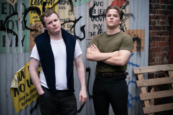 A young Phil and Grant Mitchell, portrayed by Daniel Delaney and Teddy Jay, stood against a corrugated iron backdrop covered in graffiti in a scene from EastEnders