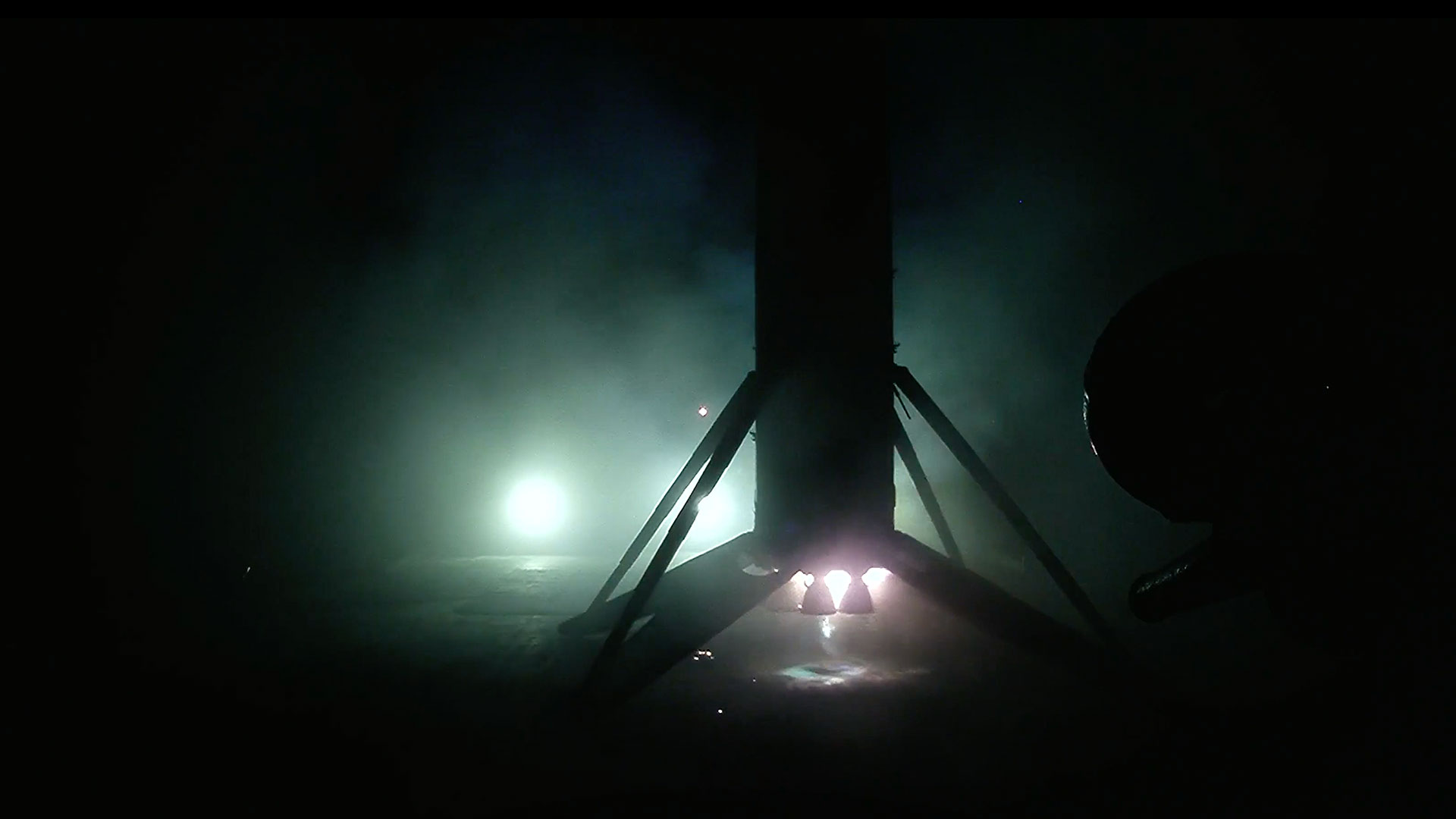the first stage of an orbital rocket is seen in silhouette, backlit by spotlights, as it stands upright on its landing legs atop an ocean-based droneship.
