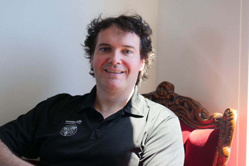 A man with dark curly hair sits on a red, velvet-cushioned armchair, smiling at the camera