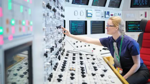 Getty Images A generic stock image of a female operator in a nuclear power station control room. She sits at a console covered in monitors and buttons and reaches to push one.