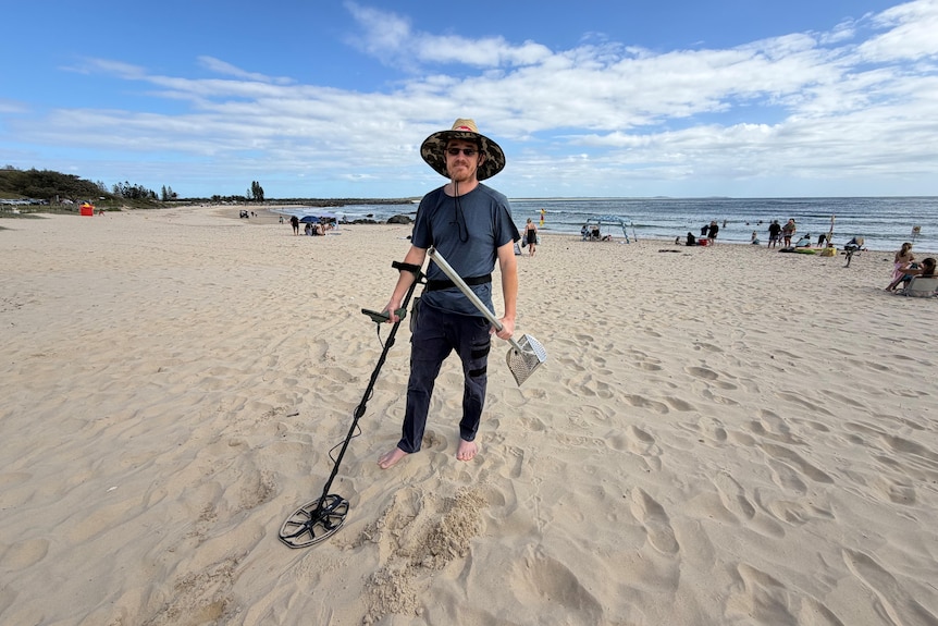 A man standing at a beach holding a metal detector and scoop.