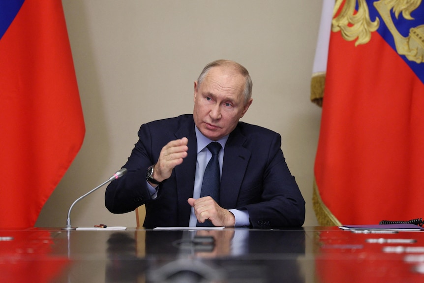 Vladimir Putin in a suit and tie sits at a table in front of Russian flags and speaks and gestures with his hands.