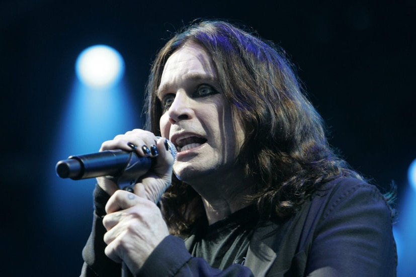 A close-up of Ozzy, with long black hair and black fingernails, holding a microphone and singing.