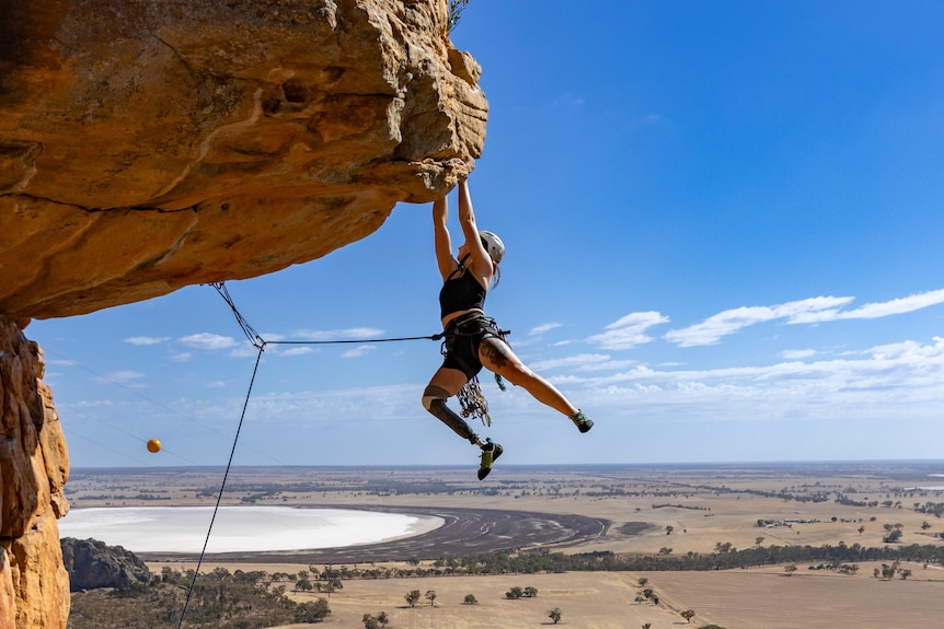 Climber Sarah Larcombe hanging by her arms off a rock wall. The Australian outback is in the background.