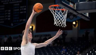 Olivier Rioux warming up before a game. He has dark hair and is wearing a white sports vest. He is holding a ball in his raised right hand as he approaches the basket to score.