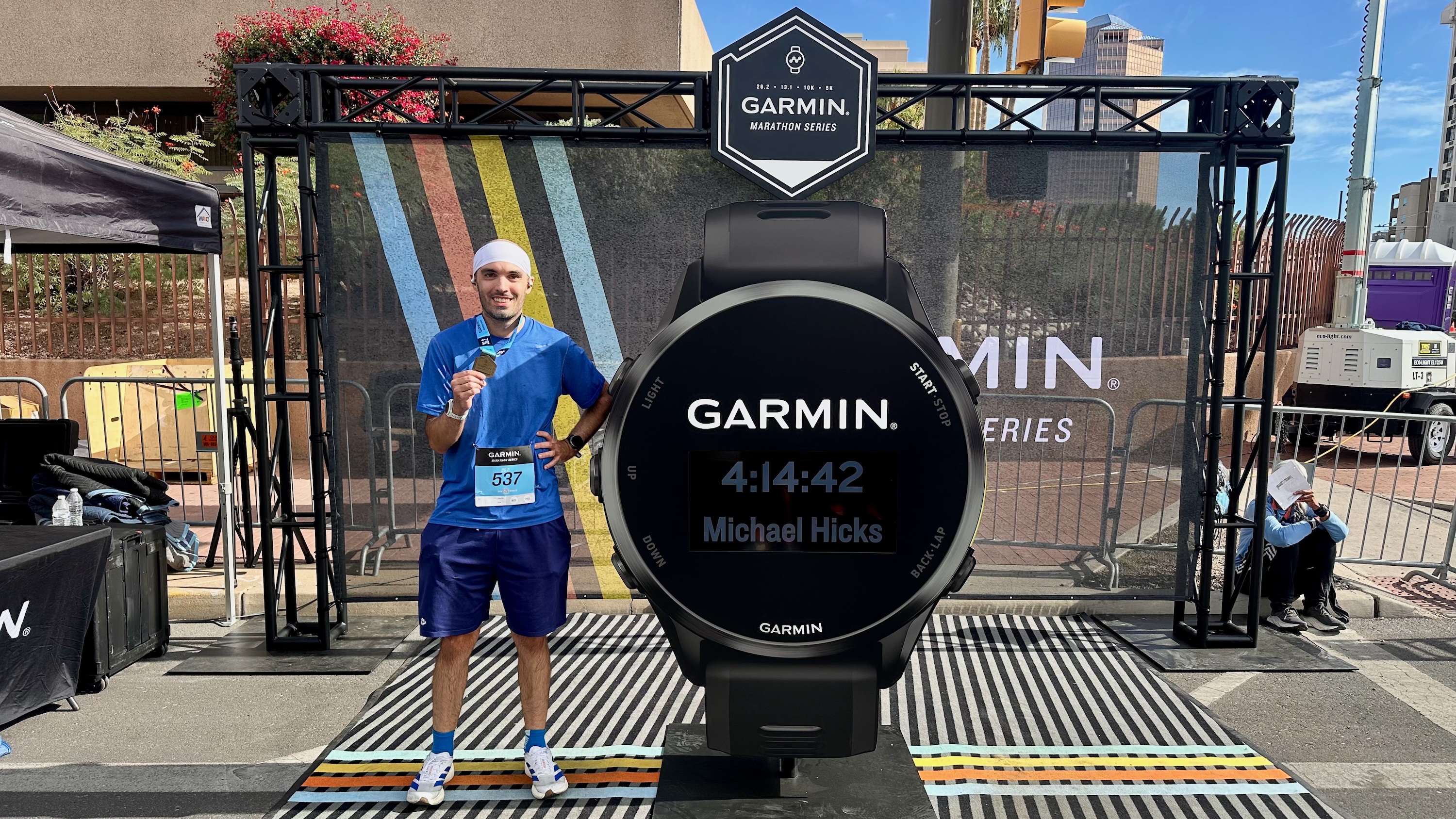 A photo of Michael Hicks standing next to a giant Garmin watch, showing his marathon time.
