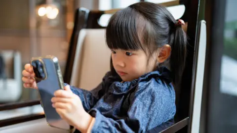 Getty Images A stock photo of little girl with pigtails looking intently at a smartphone. She is wearing a patterned blue top and sitting in a modern, warmly lit indoor room.