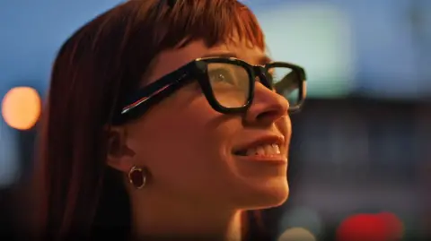 Google A close-up of a woman wearing the glasses. She is smiling and has long red hair. The glasses are black with thick rims, and the lenses seem slightly translucent.