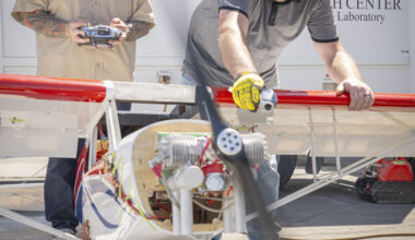 One white man with a long beard and tattoos on his arms holds a silver and gray controller, while another white man leans over to hold a small aircraft in place. The two men are both wearing hearing protection, which look like headphones. The aircraft looks to be only a few feet tall.