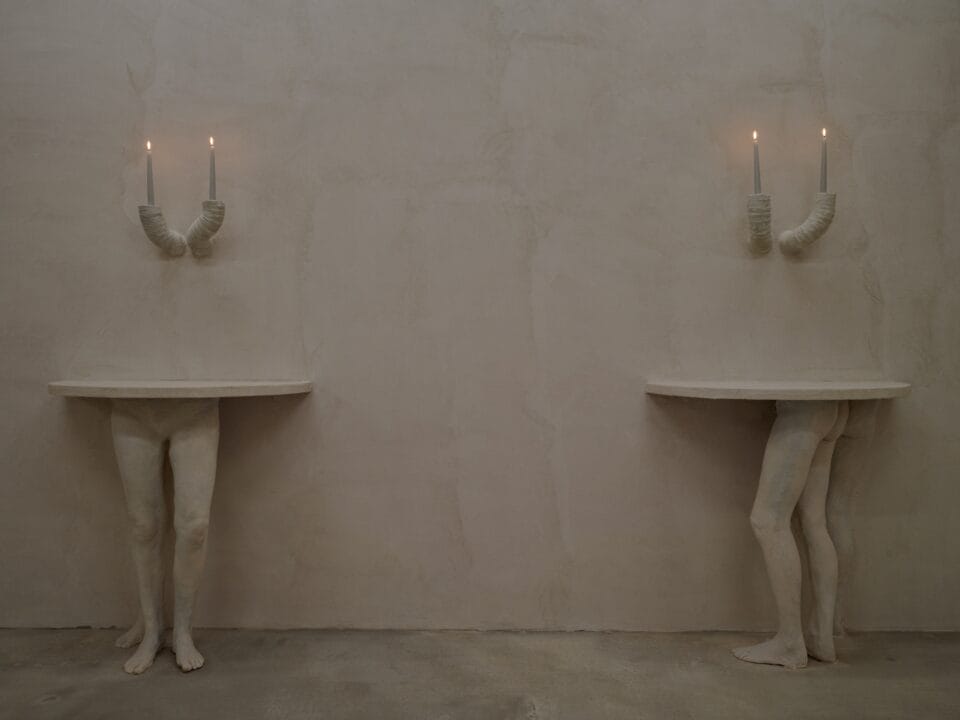 An art installation by Clementine Keith-Roach and Christopher Page of plaster-cast hands holding candles like sconces, and legs holding up two half-circle tables