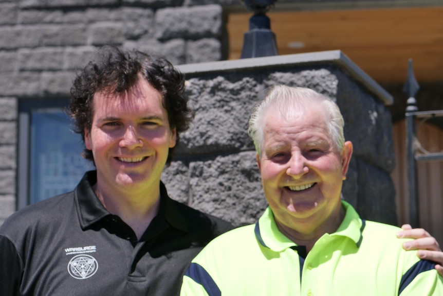 A man with dark curly hair stands with his arm around an older man with greying hair. Both smile.