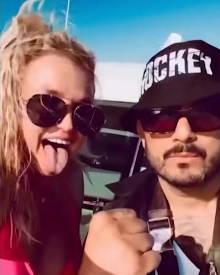 Britney Spears smiling with sunglasses and a man wearing a hockey cap on a yacht in a sunny outdoor setting.