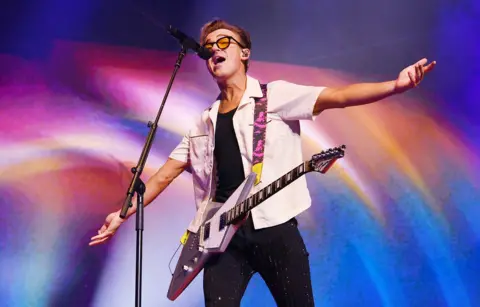 Getty Images Tom Fletcher holds his arms wide open as he sings into a microphone with a 'flying V' style guitar slung around his neck and shoulders, wearing a white shirt and black trousers against a colouful background during the Busted vs McFly tour at Wembley in September.
