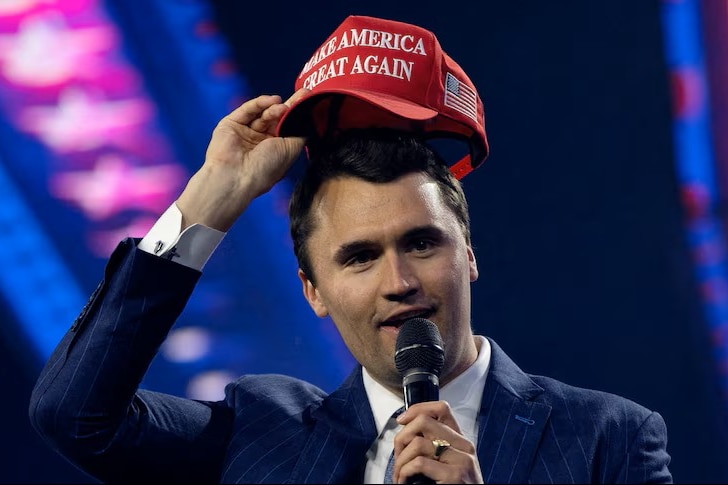 Charlie Kirk is stood with a microphone in one hand and a red cap in the other.
