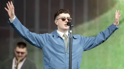 PA Media Joesef  performing onstage at an outdoor music festival - he is standing onstage and singing, while wearing a shirt, tie and blue jacket