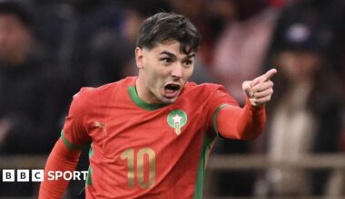 Brahim Diaz, visible from the waist up wearing a red Morocco shirt with green trim and the number 10 in gold on the front, screams in celebration and points his left hand in front of his body after scoring