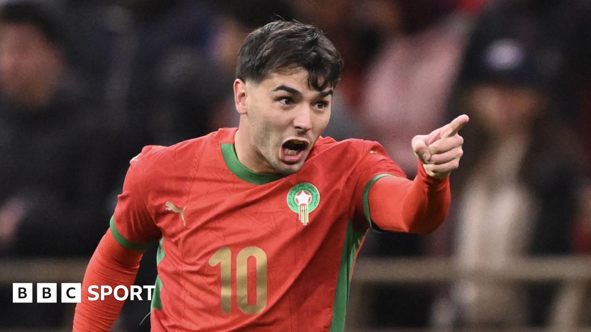 Brahim Diaz, visible from the waist up wearing a red Morocco shirt with green trim and the number 10 in gold on the front, screams in celebration and points his left hand in front of his body after scoring