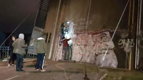 Jason Fanthorpe An image taken at night of a group of five people with ladders leaning against a disused bridge. A person at the top of the ladder is rubbing paint away from a defaced Banksy artwork as three people watch on. The deck of the bridge is in a raised position and the night sky is visible in the background.