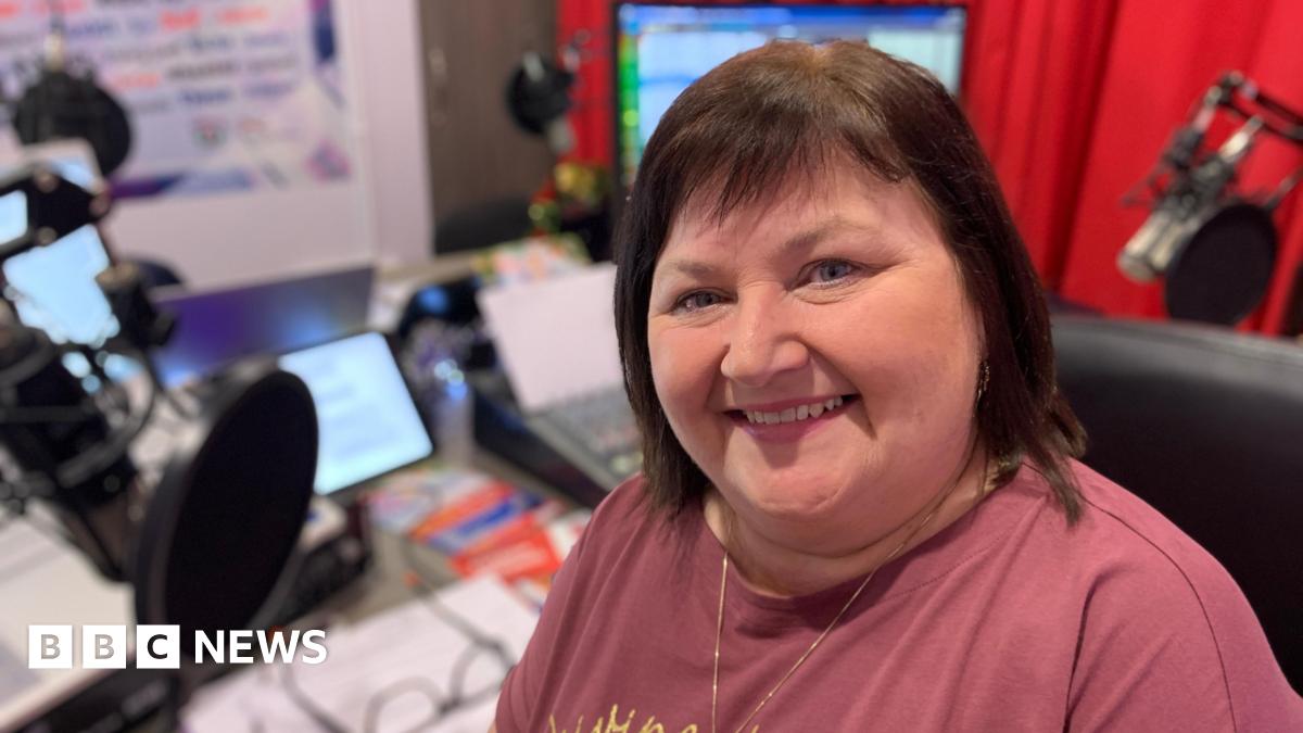 A woman with chin length brown hair is smiling at the camera. She is wearing a plum coloured t-shirt and is sitting in front of a radio studio